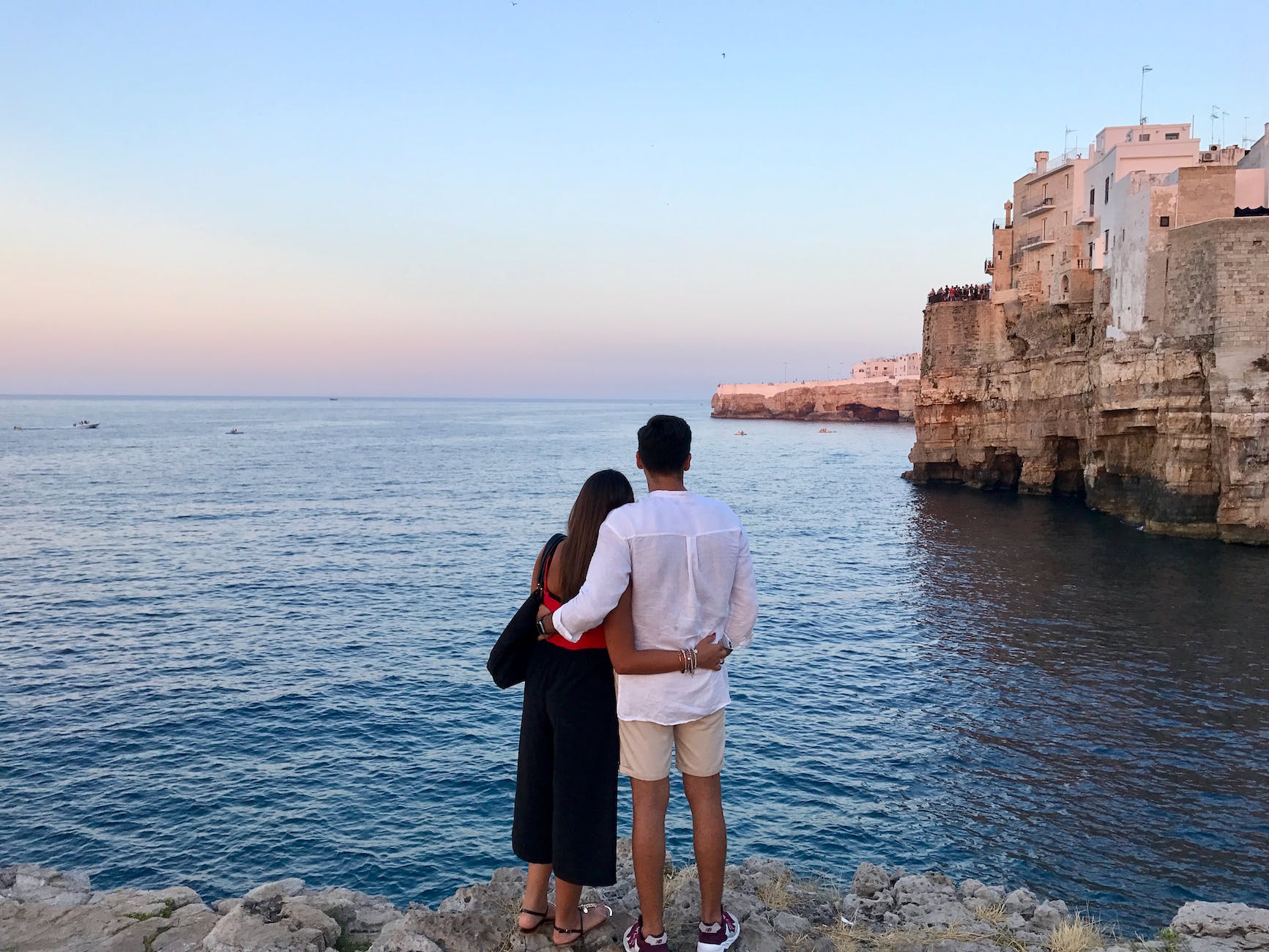 man in white dress shirt standing beside the woman in black and red dress while watching the blue calm water near brown concrete buildings under white and blue sky at daytime